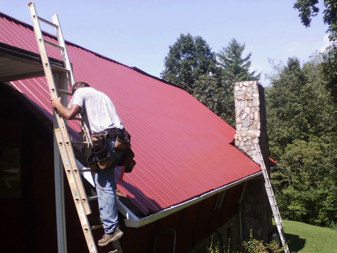 Roofer on a ladder beside a red metal roof and stone chimney — Soffit & Fascia Repair by Roofing Brothers in Prague